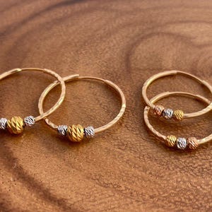 May include: Gold hoop earrings with textured detailing. Each earring features a gold, silver, and rose gold bead. The earrings are displayed on a wooden surface, showcasing their design and craftsmanship.