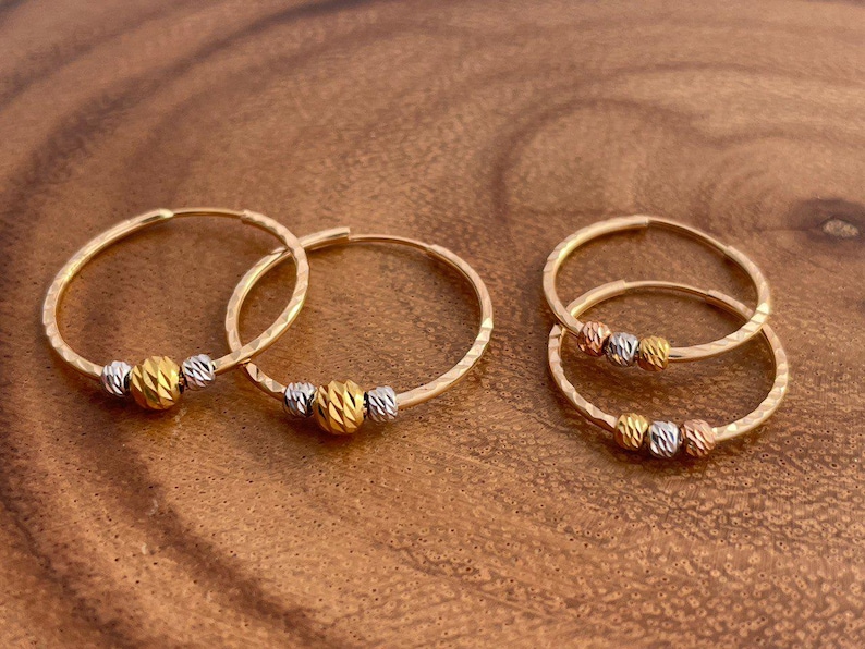 May include: Gold hoop earrings with textured detailing. Each earring features a gold, silver, and rose gold bead. The earrings are displayed on a wooden surface, showcasing their design and craftsmanship.