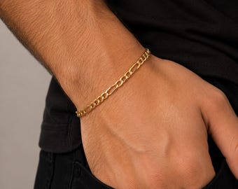May include: A gold-toned Figaro chain bracelet featuring a repeating pattern of three small circular links followed by one elongated oval link, worn around the wrist. The metal has a polished finish that reflects light, creating a bright appearance.