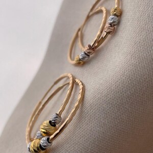 May include: Two pairs of gold hoop earrings with textured bands and three decorative, faceted beads in gold, silver, and rose gold. The earrings are displayed on a neutral, beige fabric background.