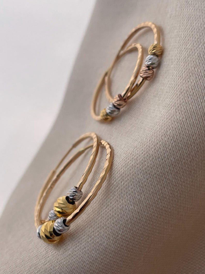May include: Two pairs of gold hoop earrings with textured bands and three decorative, faceted beads in gold, silver, and rose gold. The earrings are displayed on a neutral, beige fabric background.