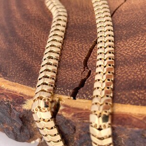 May include: A gold-colored necklace with a snake-like design. The chain is made of interlocking chevron-shaped links. The necklace is displayed on a wooden surface with a natural grain.