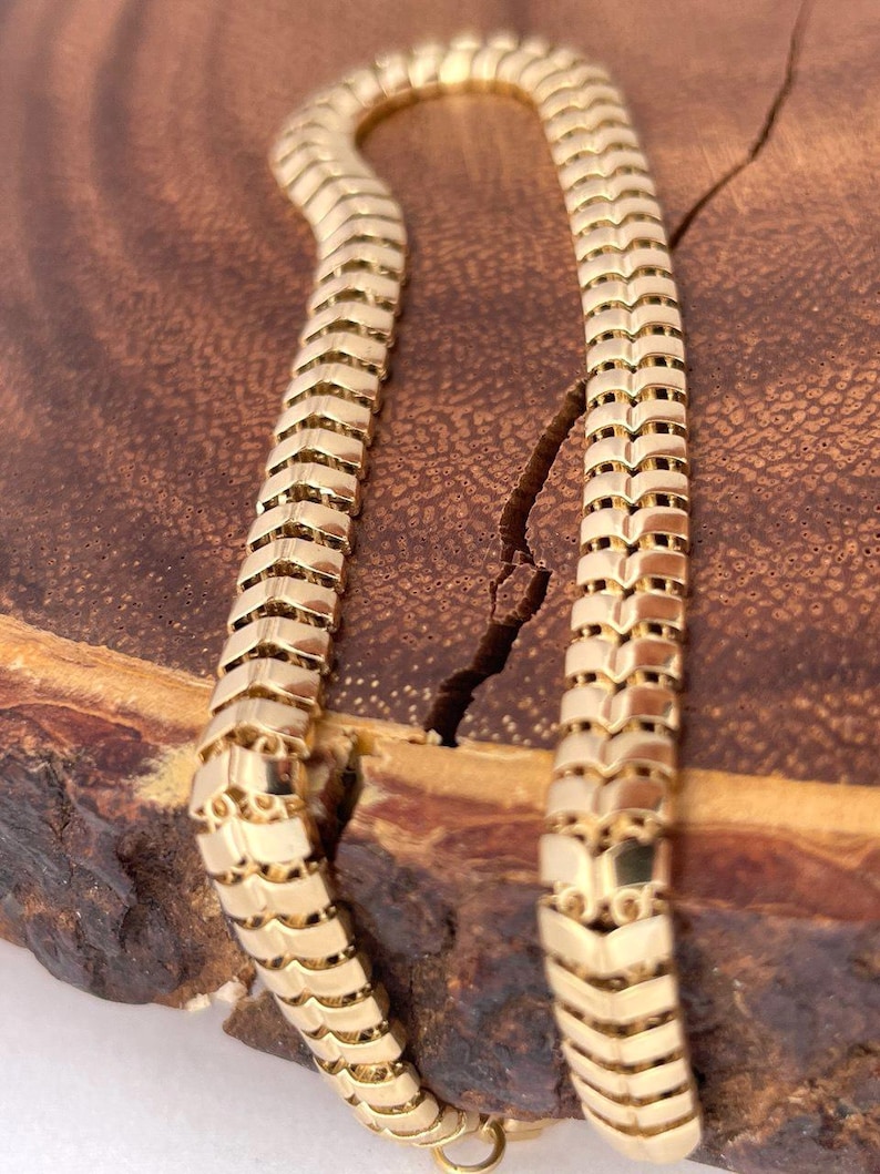 May include: A gold-colored necklace with a snake-like design. The chain is made of interlocking chevron-shaped links. The necklace is displayed on a wooden surface with a natural grain.