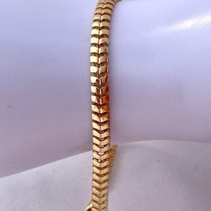 May include: A gold-colored bracelet with a chevron pattern. The bracelet is made of small, rectangular links that are connected to form a flexible band. The bracelet is approximately 7 inches in length.