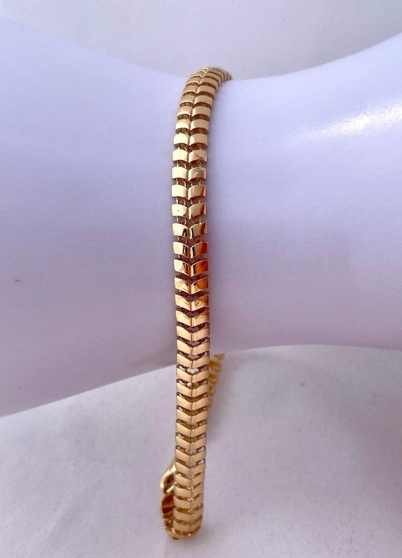 May include: A gold-colored bracelet with a chevron pattern. The bracelet is made of small, rectangular links that are connected to form a flexible band. The bracelet is approximately 7 inches in length.