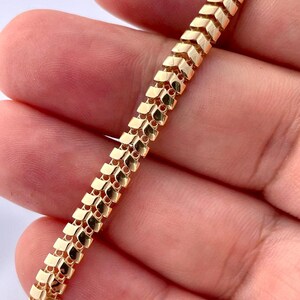 May include: A gold-colored bracelet with a unique chevron-like design. The bracelet is made of small, rectangular links that are connected to form a flexible chain. The links are arranged in a repeating pattern, creating a textured surface.