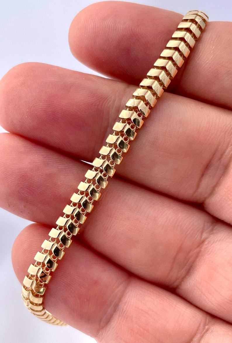 May include: A gold-colored bracelet with a unique chevron-like design. The bracelet is made of small, rectangular links that are connected to form a flexible chain. The links are arranged in a repeating pattern, creating a textured surface.