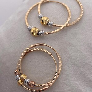 May include: Gold hoop earrings with textured surfaces and decorative beads in gold, silver, and rose gold. The earrings are displayed on a white surface. The hoops have a hammered texture.