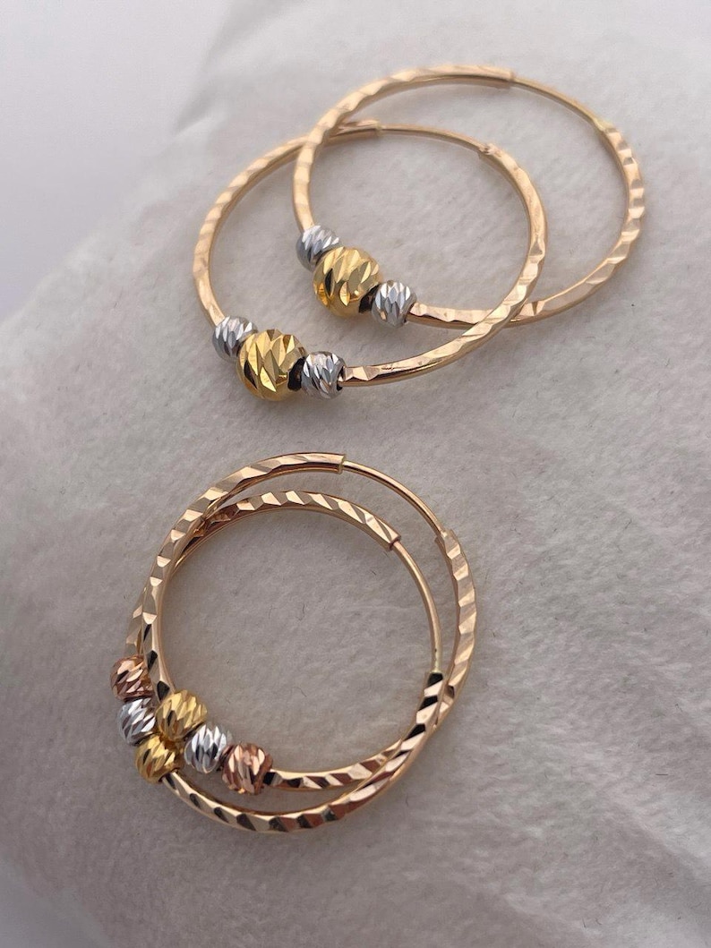 May include: Gold hoop earrings with textured surfaces and decorative beads in gold, silver, and rose gold. The earrings are displayed on a white surface. The hoops have a hammered texture.