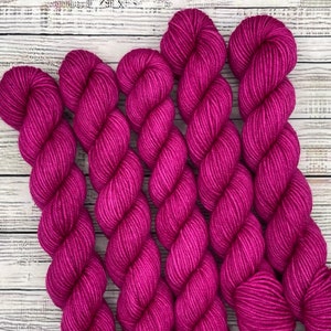 May include: Close-up of several skeins of vibrant fuchsia yarn. The yarn is tightly wound, showing the texture of the fibers. The background is a light-colored, weathered wood surface. Ideal for knitting and crochet projects.