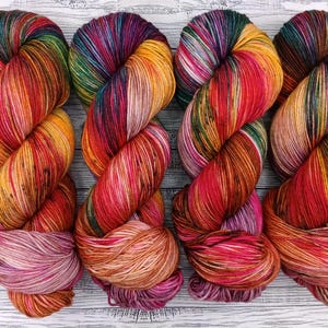 May include: Four skeins of brightly coloured yarn in shades of red, orange, yellow, and purple. The yarn is twisted, displaying the vibrant colour variations. The yarn is likely for knitting or crochet projects. Each skein is approximately 100 grams.