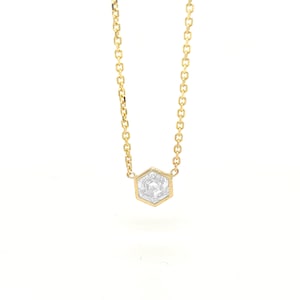 May include: A gold chain necklace featuring a single hexagonal pendant with a clear, faceted stone set in the center. The delicate metal chain connects to the top corners of the six-sided frame, creating a simple and refined jewelry piece.