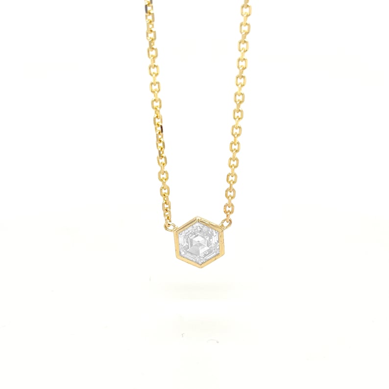 May include: A gold chain necklace featuring a single hexagonal pendant with a clear, faceted stone set in the center. The delicate metal chain connects to the top corners of the six-sided frame, creating a simple and refined jewelry piece.