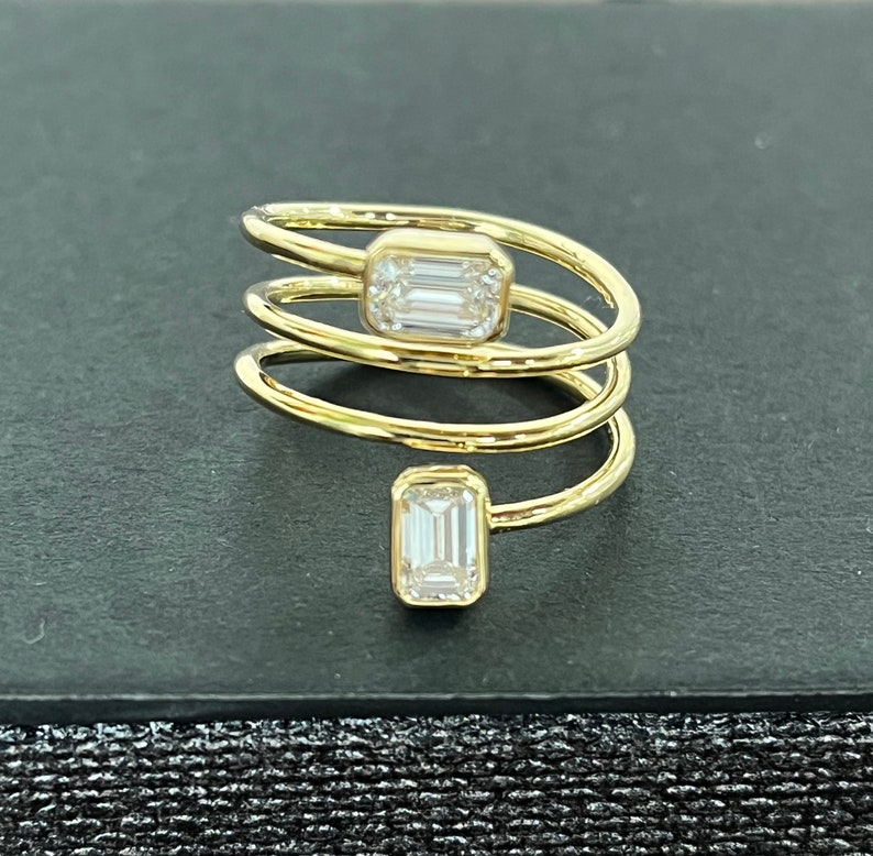 Puede incluir: Un anillo de oro con un dise&ntilde;o en espiral y dos diamantes de talla esmeralda. El anillo est&aacute; sobre un fondo negro.