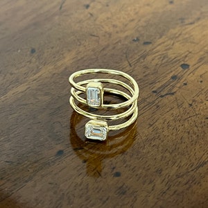 Puede incluir: Un anillo de oro con tres bandas y dos diamantes de talla esmeralda. El anillo est&aacute; sobre una superficie de madera.