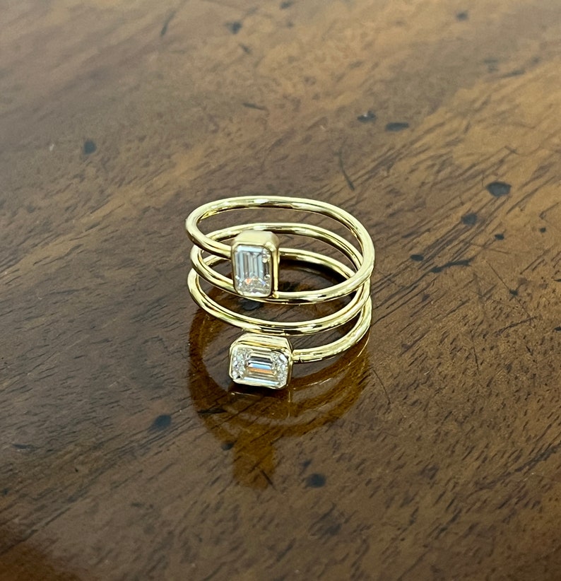 Puede incluir: Un anillo de oro con tres bandas y dos diamantes de talla esmeralda. El anillo est&aacute; sobre una superficie de madera.