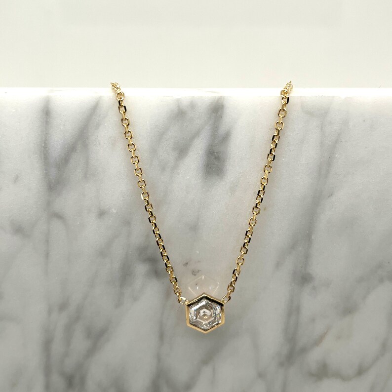 May include: A gold chain necklace featuring a single hexagonal-shaped clear gemstone pendant. The pendant is set in a gold bezel frame, and the delicate chain links are connected to the top corners of the hexagon, creating a centered focal point.