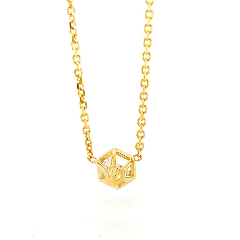 May include: A gold-toned necklace featuring a delicate chain and a central geometric pendant. The pendant is shaped like a small, open-frame cage with a hexagonal structure, enclosing a clear, faceted stone that catches the light.