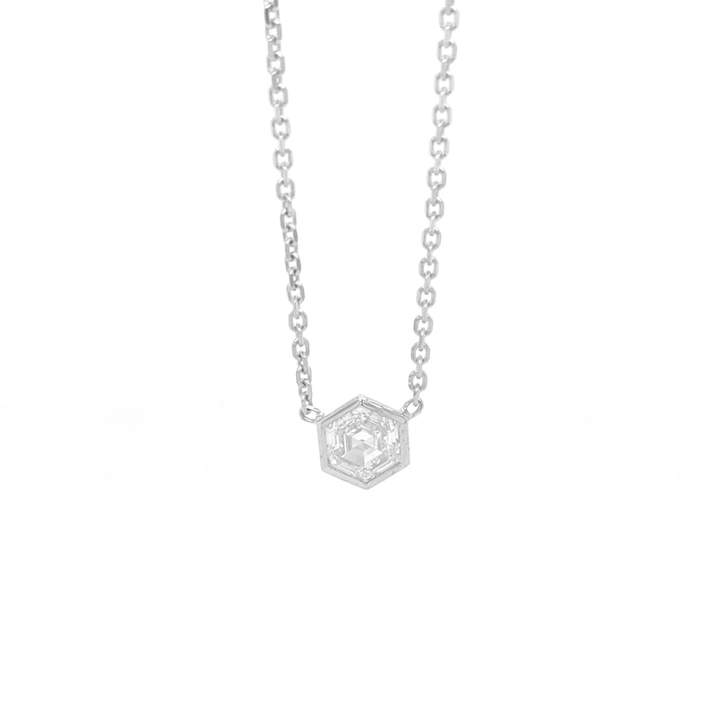 May include: A silver-toned necklace featuring a delicate chain and a single hexagonal pendant. The pendant contains a clear, faceted stone set within a metallic frame, suspended from the chain by two small loops on either side of the hexagon.