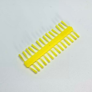 Può includere: Un pettine di plastica giallo con una fila di denti sottili e appuntiti. Il pettine è progettato per legare le mosche.