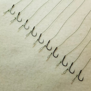 Può includere: Primo piano di un set di ami da pesca legati a fili sottili. Gli ami sono tutti neri e hanno una leggera curva. I fili sono tutti di colore verde scuro.