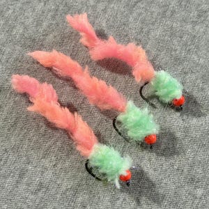 May include: Three fishing flies with pink and peach-colored bodies and light green heads. Each fly has a small red bead and a black hook. The flies are arranged on a gray fabric surface.