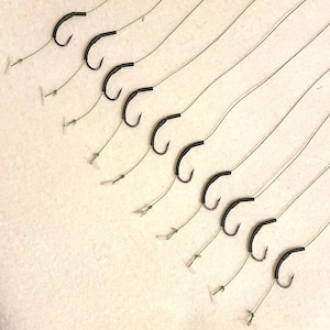 Può includere: Un set di 10 ami da pesca neri con una sottile linea trasparente attaccata a ogni amo. Gli ami sono disposti a ventaglio su una superficie bianca.