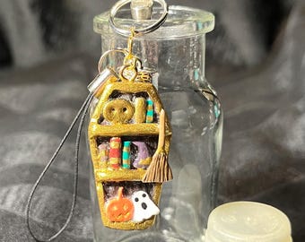 May include: A gold-colored coffin charm with a miniature bookshelf inside, a broom, a pumpkin, and a ghost. The charm is attached to a clear glass bottle with a clear plastic lid.
