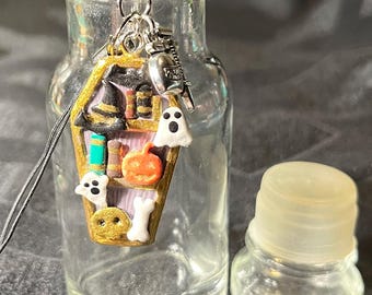May include: A clear glass bottle with a cork stopper and a charm attached to the lid. The charm is a small, gold-colored coffin with a witch's hat, a ghost, a pumpkin, and bones on it. The bottle is sitting on a dark gray surface.