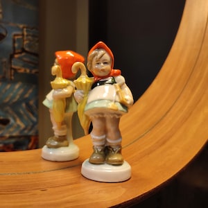 Puede incluir: Una figurita de porcelana de una niña que lleva un vestido rojo y blanco y sostiene un paraguas. La figurita está de pie sobre una base blanca.