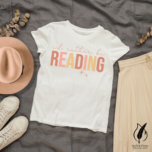 Peut inclure: T-shirt blanc avec le texte "i'd rather be READING" en lettres roses, orange et jaunes. Le texte est dans un style dégradé avec un design de paillettes.