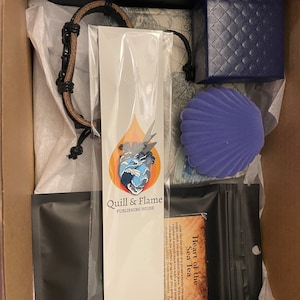 Op de afbeelding: Een doos gevuld met verschillende items, waaronder een zwarte leren armband, een blauw en wit hartvormige pin, een paarse fluwelen schelp, een blauwe fluwelen doos en een zwarte tas met een wit label waarop "Heart of the Sea Tea" staat. Het label bevat ook een beschrijving van de thee. De doos bevat ook een witte kaart met de tekst "Quill & Flame Publishing House".
