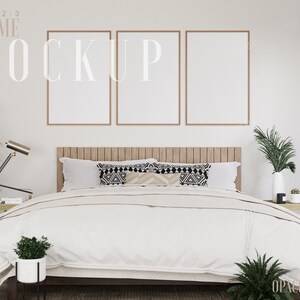 May include: Three empty picture frames with a 2:3 aspect ratio, hanging on a white wall above a bed with white bedding and patterned pillows. The bed is in a bedroom with a wooden headboard and a nightstand with a plant and a lamp.