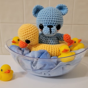 May include: A crocheted blue bear and a yellow duck float in a clear bowl with blue fabric. Several yellow rubber duckies surround the duck and bear. The bear has black eyes and an orange nose. The duck has black eyes and an orange beak.