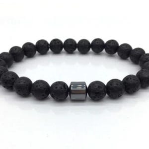 May include: A black lava stone bracelet with a rectangular, metallic accent bead. The bracelet features round, porous beads strung together, creating a simple yet stylish accessory. The bracelet is displayed on a white background.