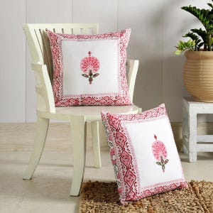 May include: Two white cotton cushion covers with pink floral designs. The cushion covers have a white background with a pink border and a pink floral design in the centre. The cushion covers are sitting on a white chair and a brown rug.