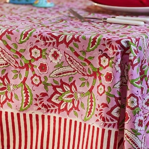 May include: A pink floral tablecloth with a red and green botanical design. The tablecloth has a border of red and white vertical stripes. The table is set with red napkins, plates, and silverware.