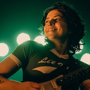 May include: A woman with short, curly brown hair smiles while playing an electric guitar. She is wearing a black and white striped shirt with the word "Lie" on it. The background is a dark green with white lights.