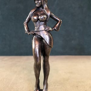 May include: A metallic bronze-colored figurine of a standing person with long hair, wearing a fitted top with long sleeves and a short skirt. The figure has hands placed on the hips and is mounted on a small, circular, matching bronze-colored base.