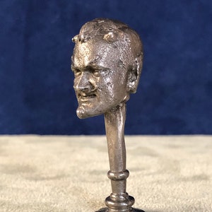 May include: A silver metal sculpture of a man's head with horns, mounted on a pedestal. The sculpture is detailed and realistic, with a textured surface.