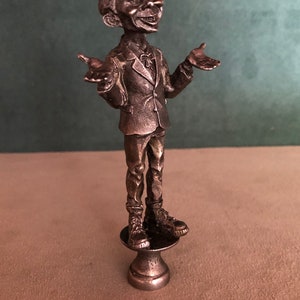May include: A silver-toned figurine of a man in a suit, with arms outstretched. The figure stands on a small pedestal. The man has a smiling expression and detailed facial features. The figurine is likely made of metal.