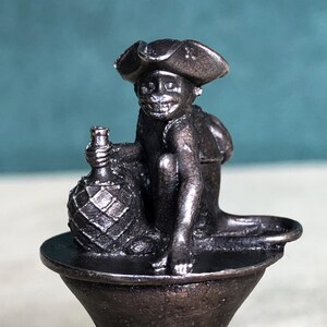 May include: A silver metal bottle stopper featuring a monkey wearing a tricorn hat and holding a bottle. The monkey is sitting on a round base.