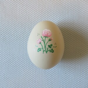 May include: A cream-colored, egg-shaped decorative item. It features a hand-painted floral design with pink flowers, green stems, and leaves. The egg is a decorative item, possibly for Easter or spring.