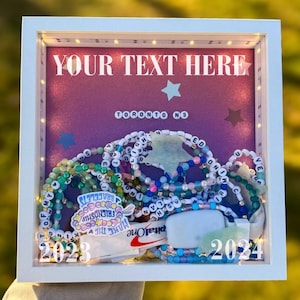 Può includere: Una scatola di ombra bianca con uno sfondo rosa e luci bianche. La scatola contiene una varietà di braccialetti di perline colorate, una saponetta bianca e un adesivo che dice "Make the Friendship Bracelet". Il testo "YOUR TEXT HERE" è nella parte superiore della scatola, e il testo "TORONTO N3" è al centro della scatola. I numeri "2023" e "2024" sono nella parte inferiore della scatola.