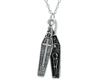 May include: Silver-toned necklace featuring two coffin pendants. One coffin has a cross design, the other a skeleton. The pendants are connected to a delicate chain, creating a gothic or Halloween-themed accessory.