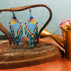 Pu&ograve; includere: Un paio di orecchini con perline blu e dettagli floreali colorati appesi a un ferro da stiro vintage. Gli orecchini sono esposti su una superficie rustica con un annaffiatoio dorato pieno di fiori rosa.