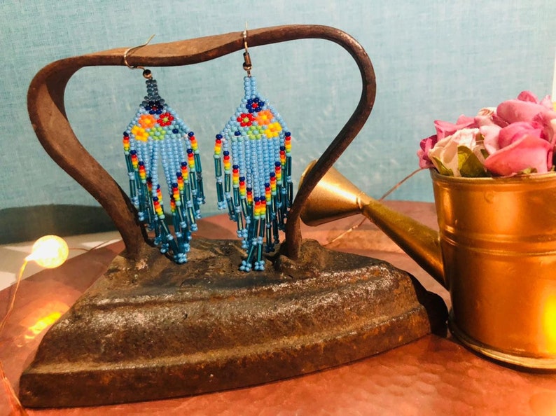 Pu&ograve; includere: Un paio di orecchini con perline blu e dettagli floreali colorati appesi a un ferro da stiro vintage. Gli orecchini sono esposti su una superficie rustica con un annaffiatoio dorato pieno di fiori rosa.
