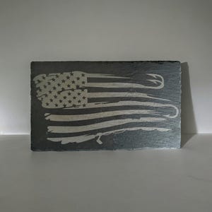 May include: A rectangular dark gray slate plaque featuring a laser-engraved design of the American flag. The stripes of the flag are stylized to incorporate the shape of a fishing hook and line, with the stars visible in the upper left corner.