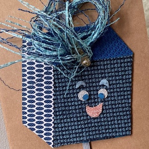 May include: A blue and white patterned house-shaped tag with a smiling face. The tag has a blue and white fringe on top.