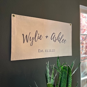 May include: A beige canvas banner with the text "Wylie + Ashlee" in a cursive font and "Est. 11.11.22" in a smaller font below. The banner is hanging on a dark green wall.